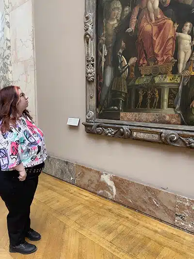 A photo of Ashley Couto staring at a painting by Andrea Mantegna in The Louvre.